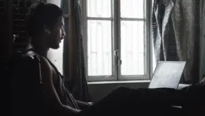 Parent reading on a laptop beside a sunny window with sheer curtains